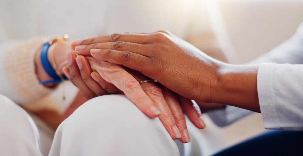 Two people are gently holding hands, conveying support and empathy. The scene is close-up, focusing on their hands and wrists.