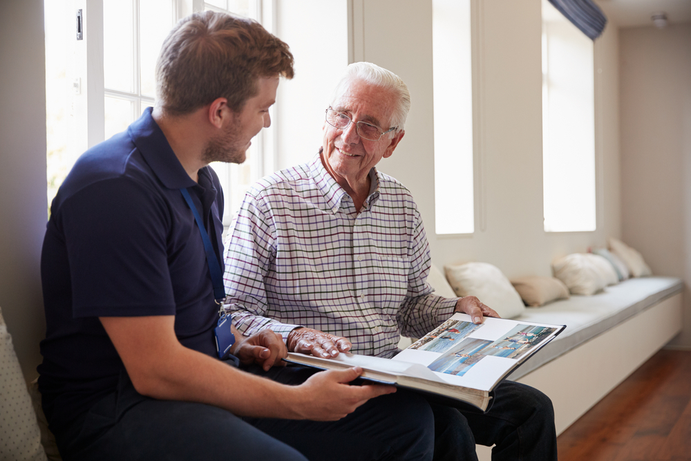 Two people sit by a window, sharing a photo album and smiling. The setting is cozy with cushions along the bench.