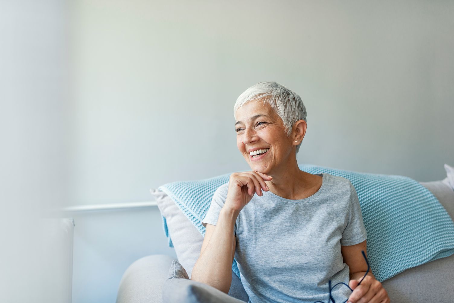 A person with short grey hair sits on a couch, smiling and holding glasses, with a blue blanket draped behind them.