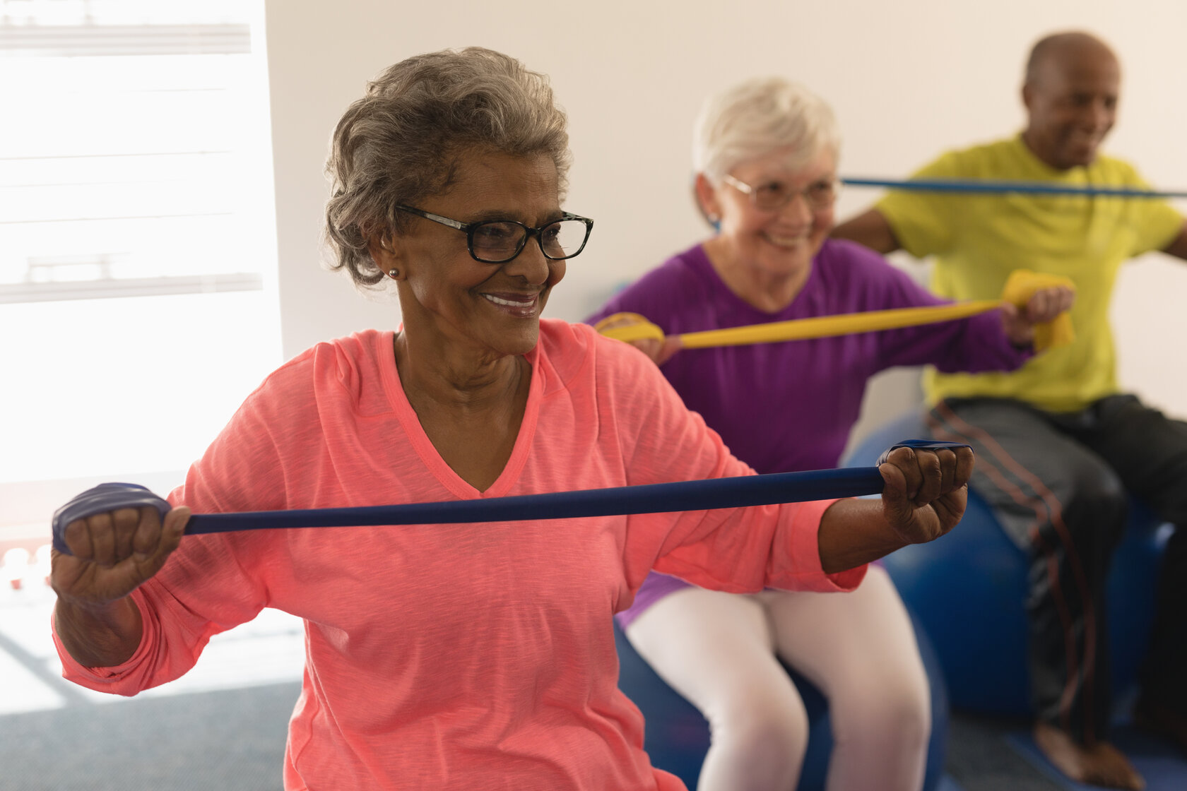 Three people are exercising with resistance bands, sitting on stability balls, smiling, and appearing engaged in a bright indoor fitness setting.