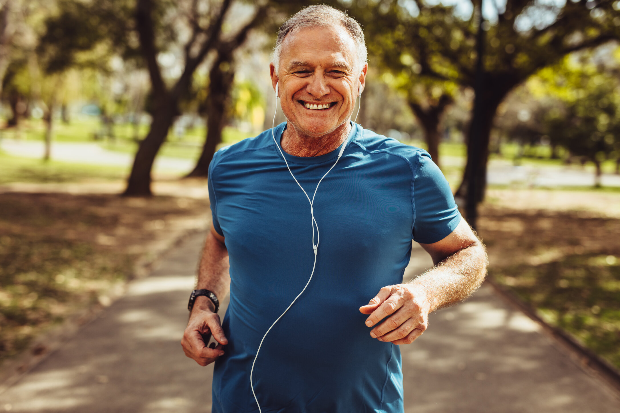 A person in a blue shirt jogs along a tree-lined park path, wearing earphones, on a sunny day.