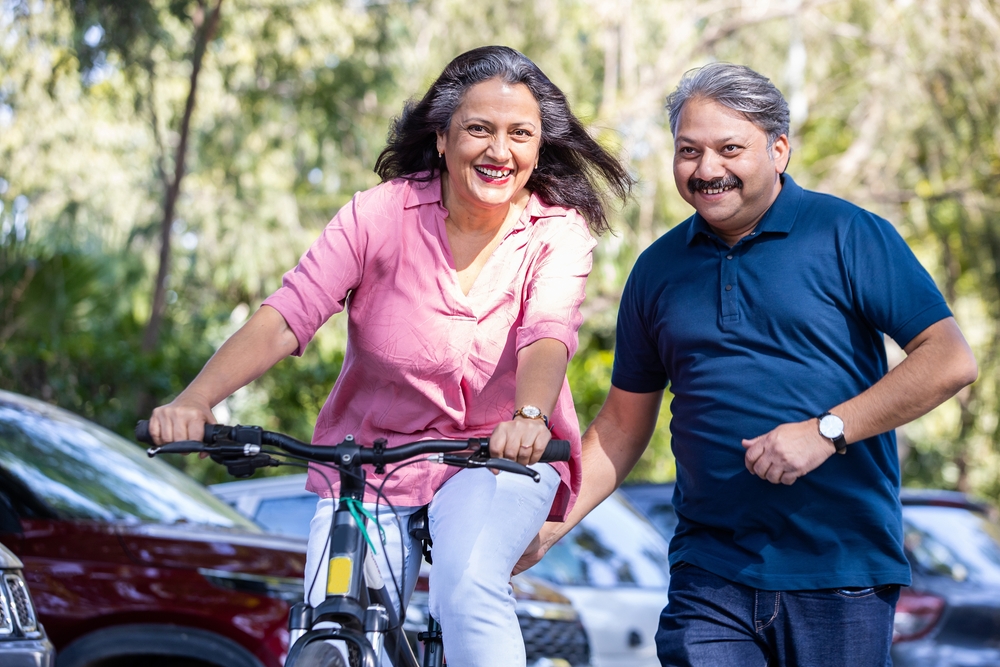 A person rides a bicycle while another person walks beside them, both smiling, outdoors with trees and parked cars in the background.