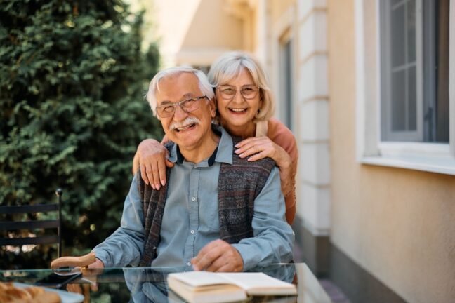 Two smiling people sitting outdoors at a table with a book. There's greenery in the background and a building with windows.