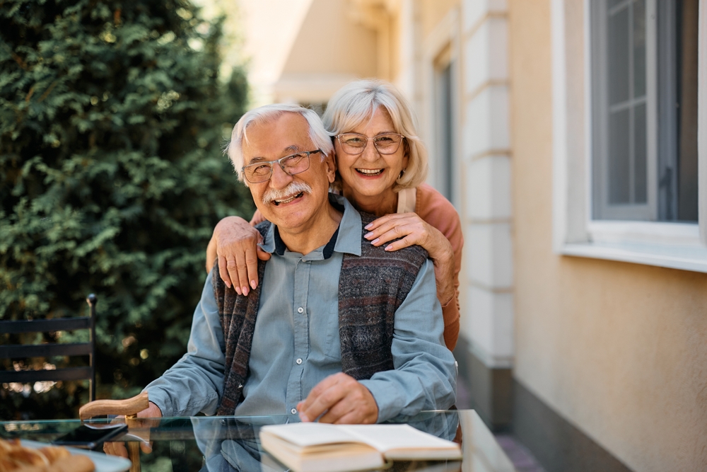 Two smiling people sitting outdoors at a table with a book. There's greenery in the background and a building with windows.