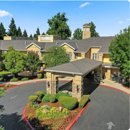 A two-story building with a covered entrance and landscaped garden, surrounded by trees, under a clear blue sky. No recognizable landmarks are visible.