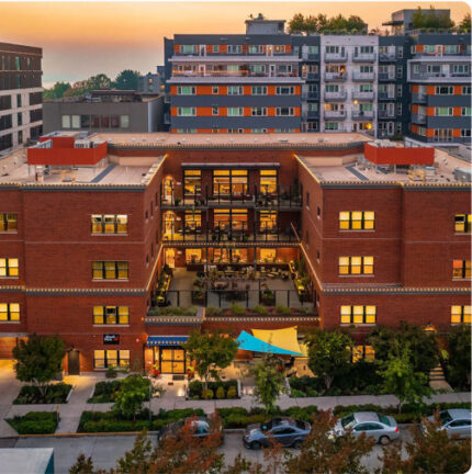 Modern urban buildings at sunset, featuring a red brick structure with lit windows. Cars parked on the street, trees lining the sidewalk.