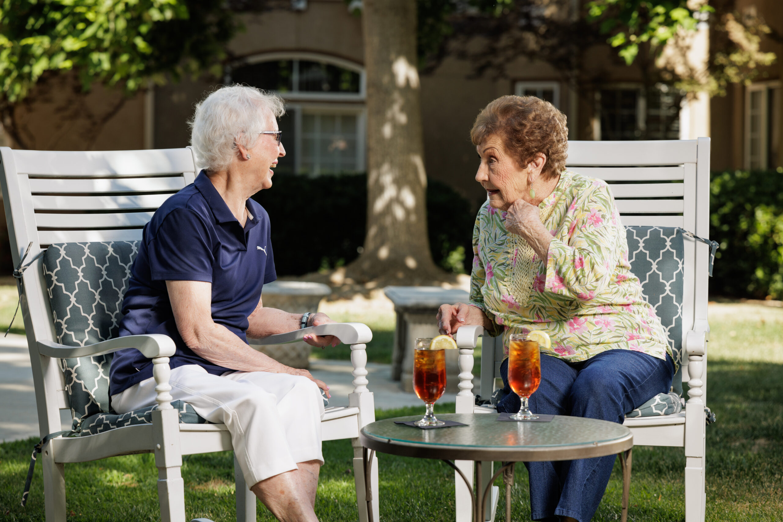 Two people chatting outdoors, seated on chairs, with iced drinks on a table. Surrounded by trees and a building in the background.