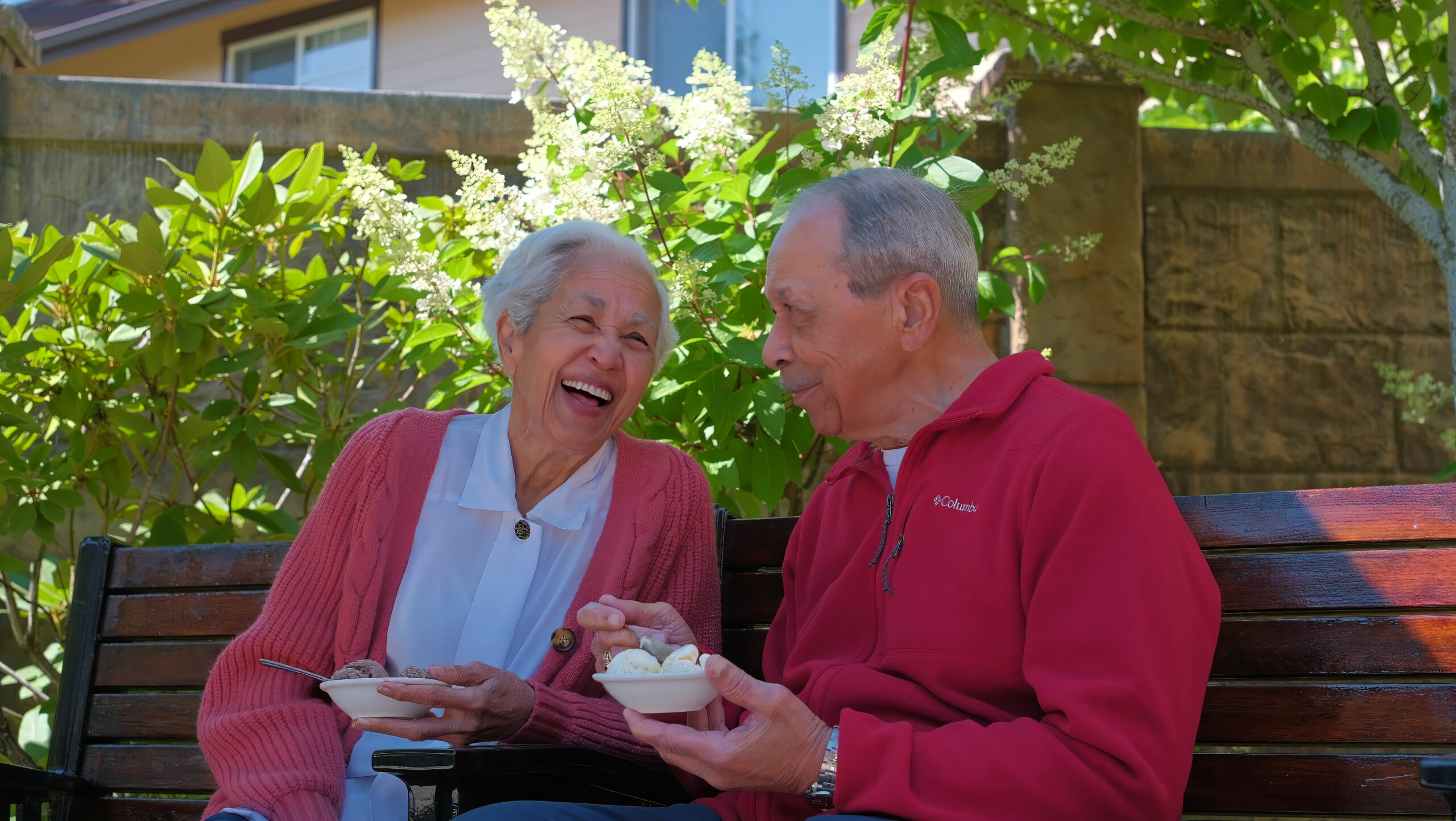 Two people sitting on a bench, laughing and enjoying ice cream in a sunny garden setting with blooming flowers.