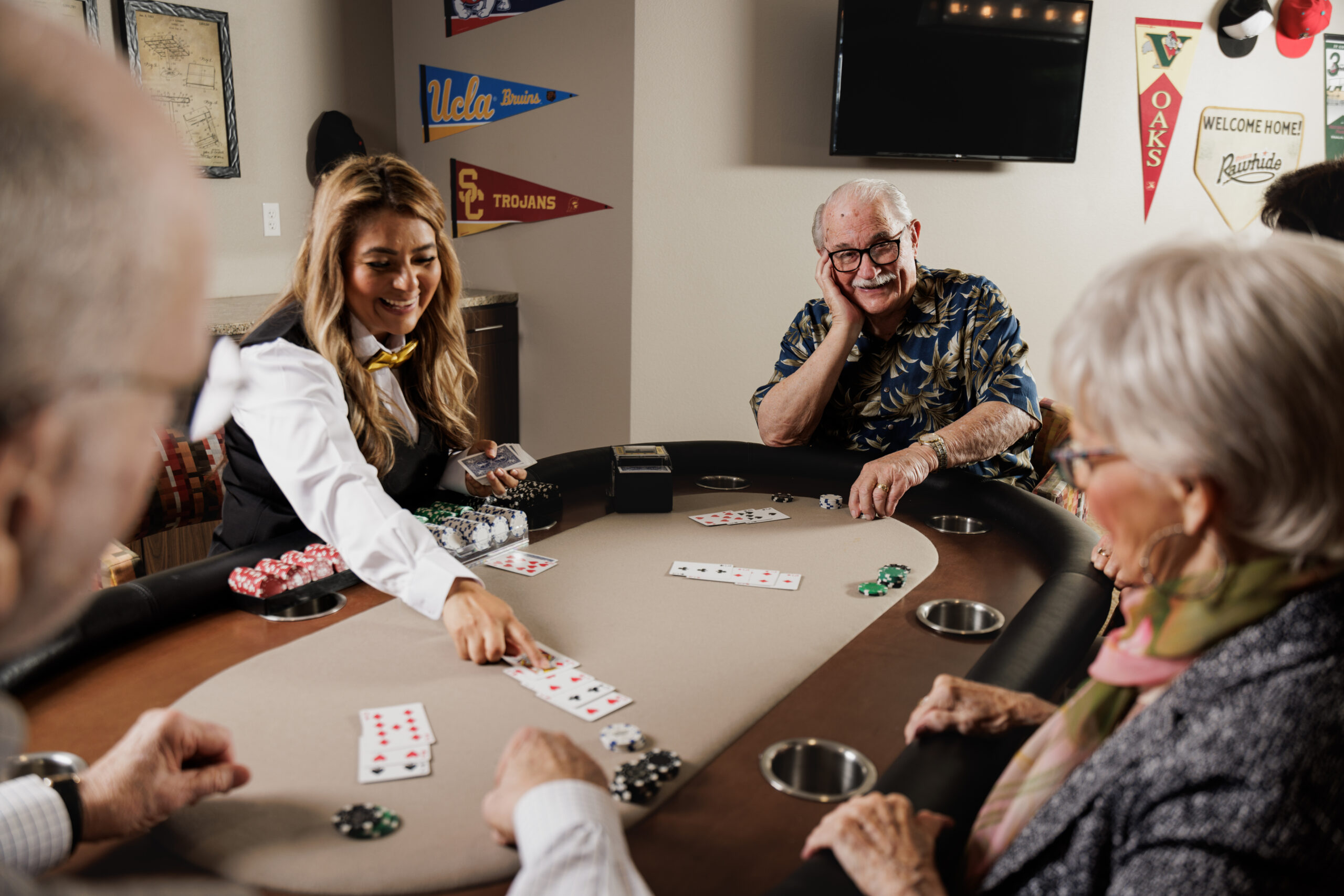 People play poker around a table in a room with university pennants. A person wearing a vest deals cards, engaging in the game.