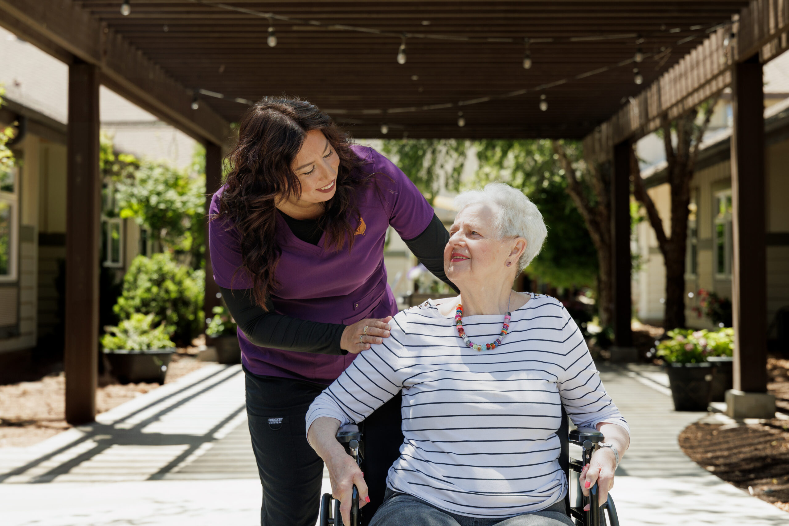 A person in purple assists another in a wheelchair under a wooden pergola in a sunny, landscaped garden setting.