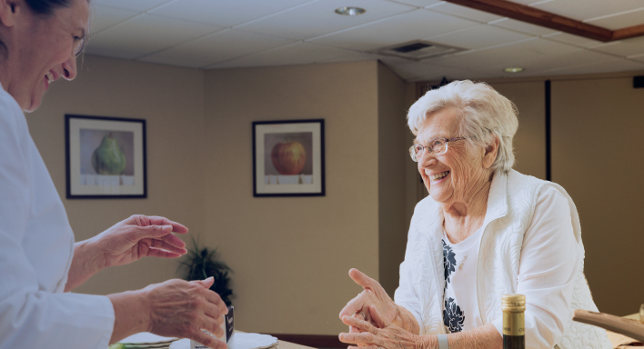 Two people smiling and conversing indoors, with framed fruit artwork on the wall. A bottle is visible on the table.