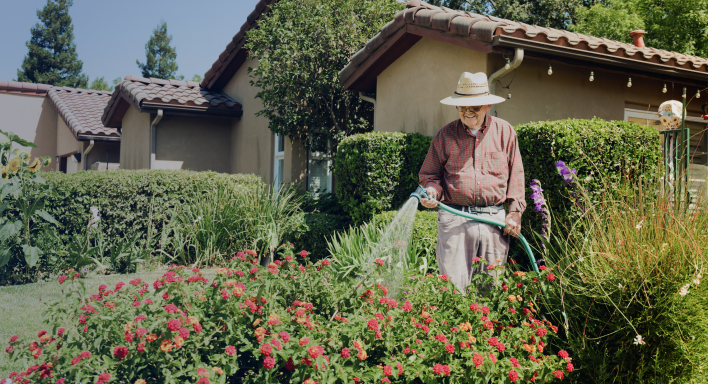 A person in a straw hat waters a lush garden with red flowers, standing in front of a house with tiled roof.