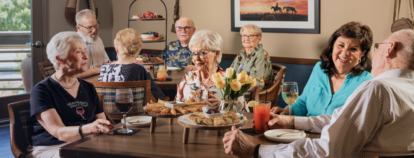 A group of people enjoy food and drinks around a table indoors, with a painting and flowers in the background, creating a cheerful atmosphere.