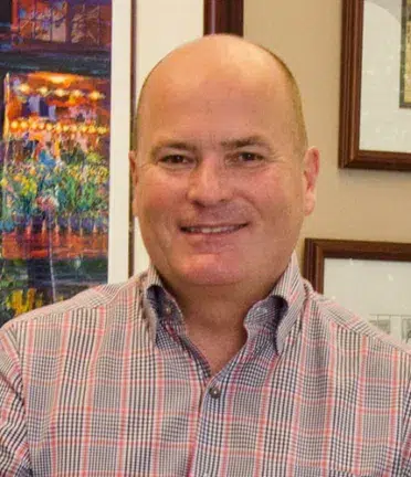 A bald person with a collared shirt, standing inside against a wall with picture frames. No landmarks visible.