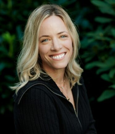 A person with short blonde hair smiles, wearing a black top, set against a leafy green background. The image is closely cropped, showing upper body only.