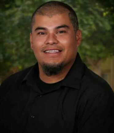 A person with short hair and a goatee smiles, wearing a black shirt. The background features green, blurred foliage.