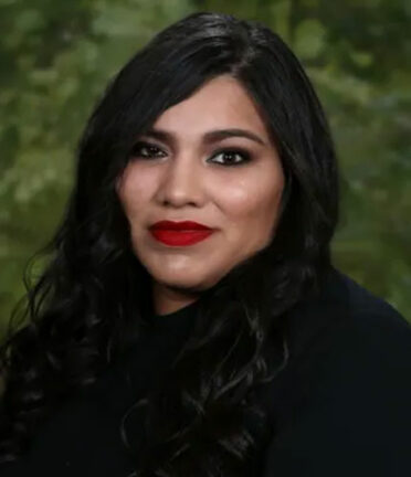 A person wearing a black sweater, long hair with red lipstick smiling against a blurred-forest background. There are no recognizable landmarks or historical buildings.