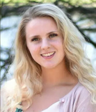 A person with long, wavy blonde hair with head tilted to one side, wearing a pink shirt stands outdoors, smiling at the camera.