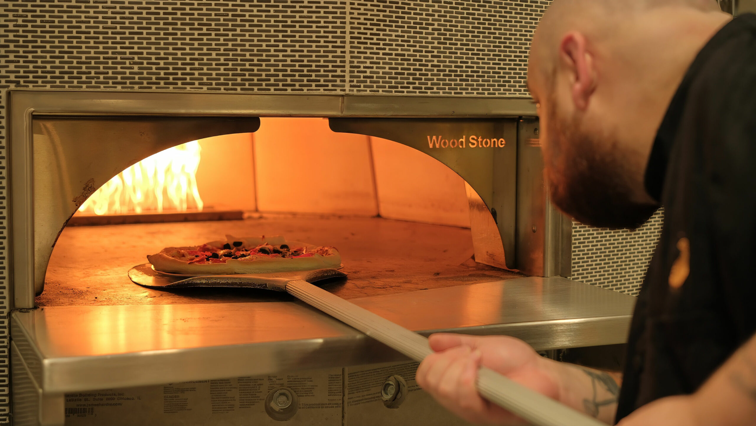 A person uses a peel to place a pizza into a Wood Stone oven with visible flames, suggesting a professional kitchen setting.