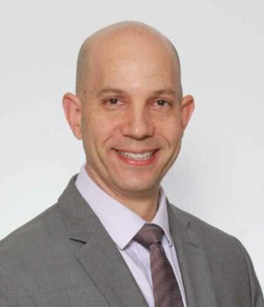 A person wearing a gray suit and tie is smiling against a plain white background. There are no recognizable landmarks or historical buildings.