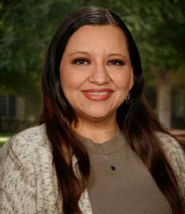 A person smiling, standing outdoors with trees in the background. No recognizable landmarks or historical buildings are visible.