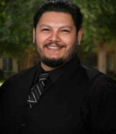 A person with a beard and dark clothing smiles outdoors, with trees and a building blurred in the background. No landmarks or historical buildings visible.