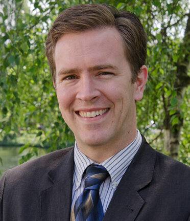 A person wearing a gray suit and tie is smiling outside in front of a large tree. There are no recognizable landmarks or historical buildings.