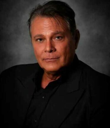 A person with short dark hair and a black suit is posing against a dark, neutral background. The focus is on their face and expression.