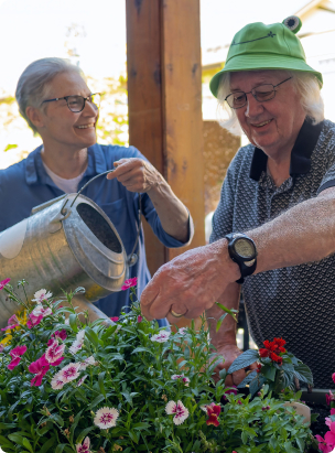 Two people happily tend to colorful flowers. One person waters them while the other prunes. They appear to be in a garden setting.