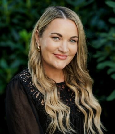 A person with blonde, wavy hair smiles, wearing a black top, set against a leafy green background. The image is closely cropped, showing upper body only.