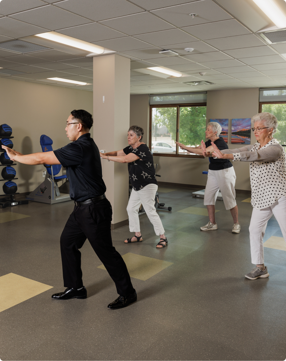 Four people practicing Tai Chi in a well-lit room with exercise equipment and scenic photographs on the walls.