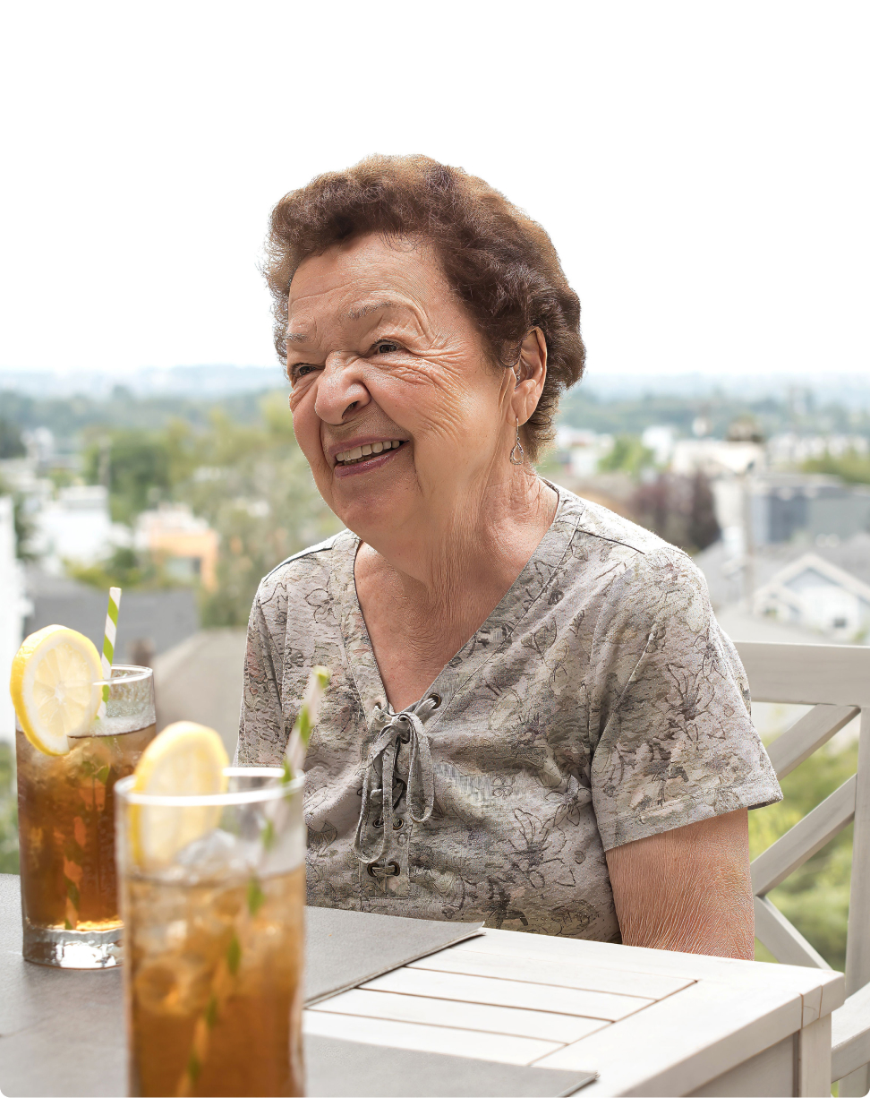 A person smiling, seated outdoors with two iced tea glasses on a table, overlooking a blurry urban landscape in the background.