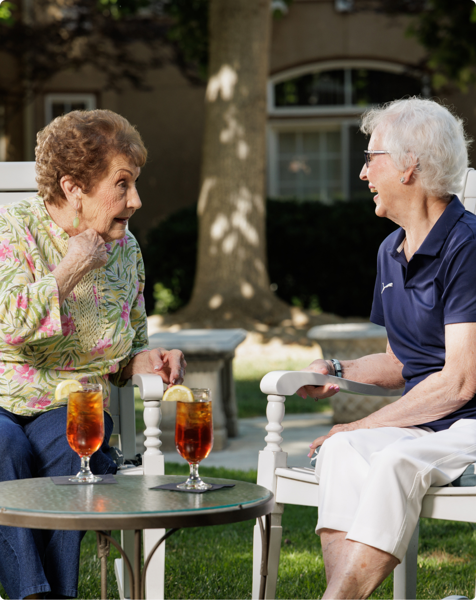 Two people sitting outdoors, enjoying iced tea and conversing. Surrounded by trees and a building in the background, creating a relaxed atmosphere.