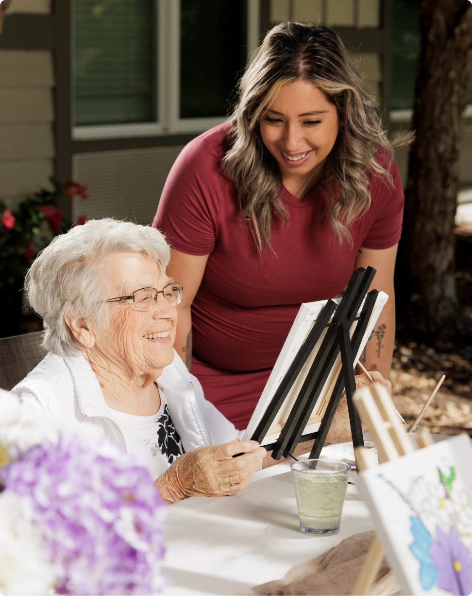 Two people enjoying painting together outside. One is painting on a canvas while the other looks on, smiling. Flowers and drink nearby.