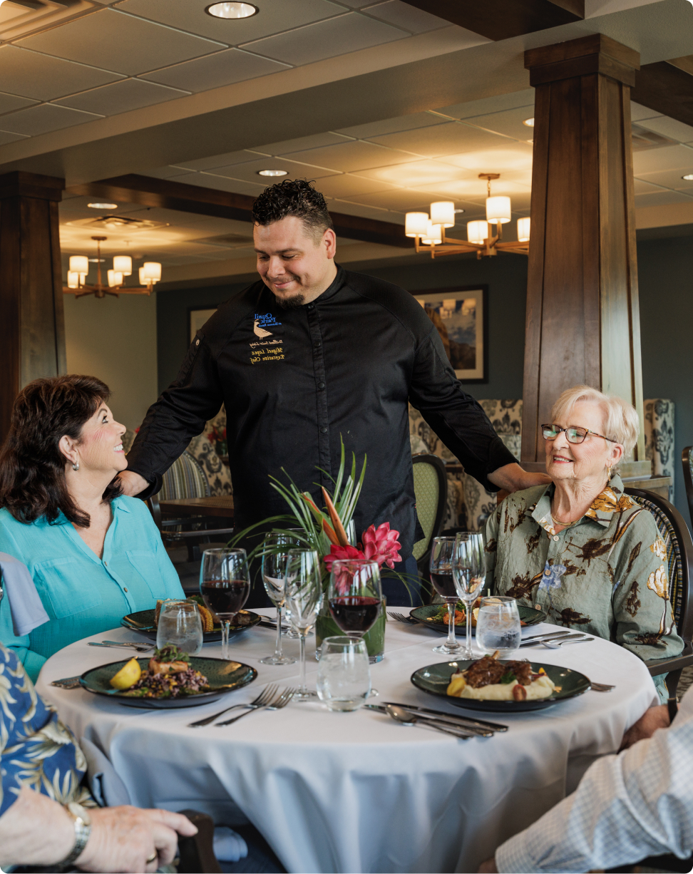 A person in a black chef uniform interacts with three people seated at a round table adorned with food and flowers in a restaurant.