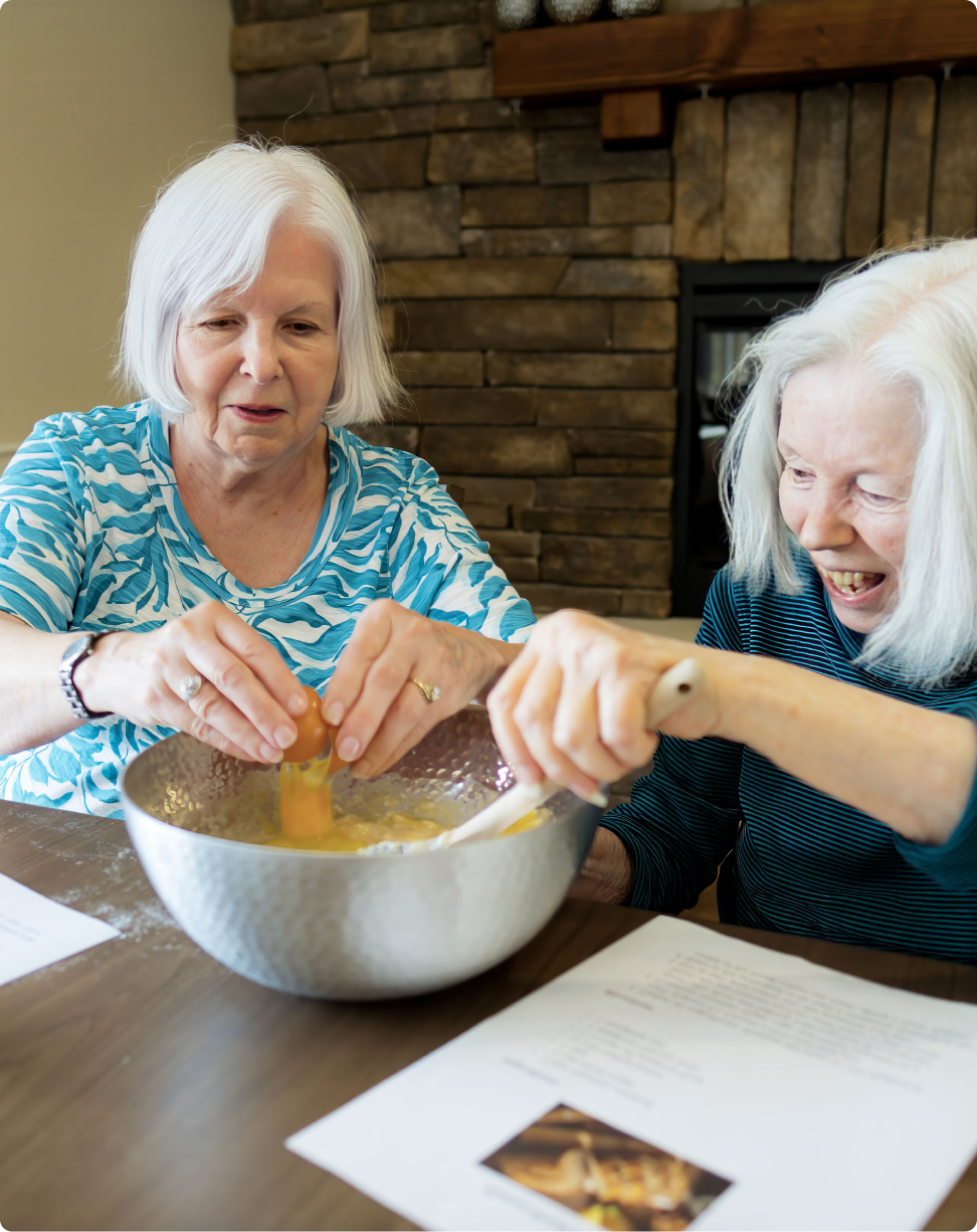 Two people are joyfully baking together, cracking eggs into a bowl, with recipe sheets on the table and a stone fireplace in the background.