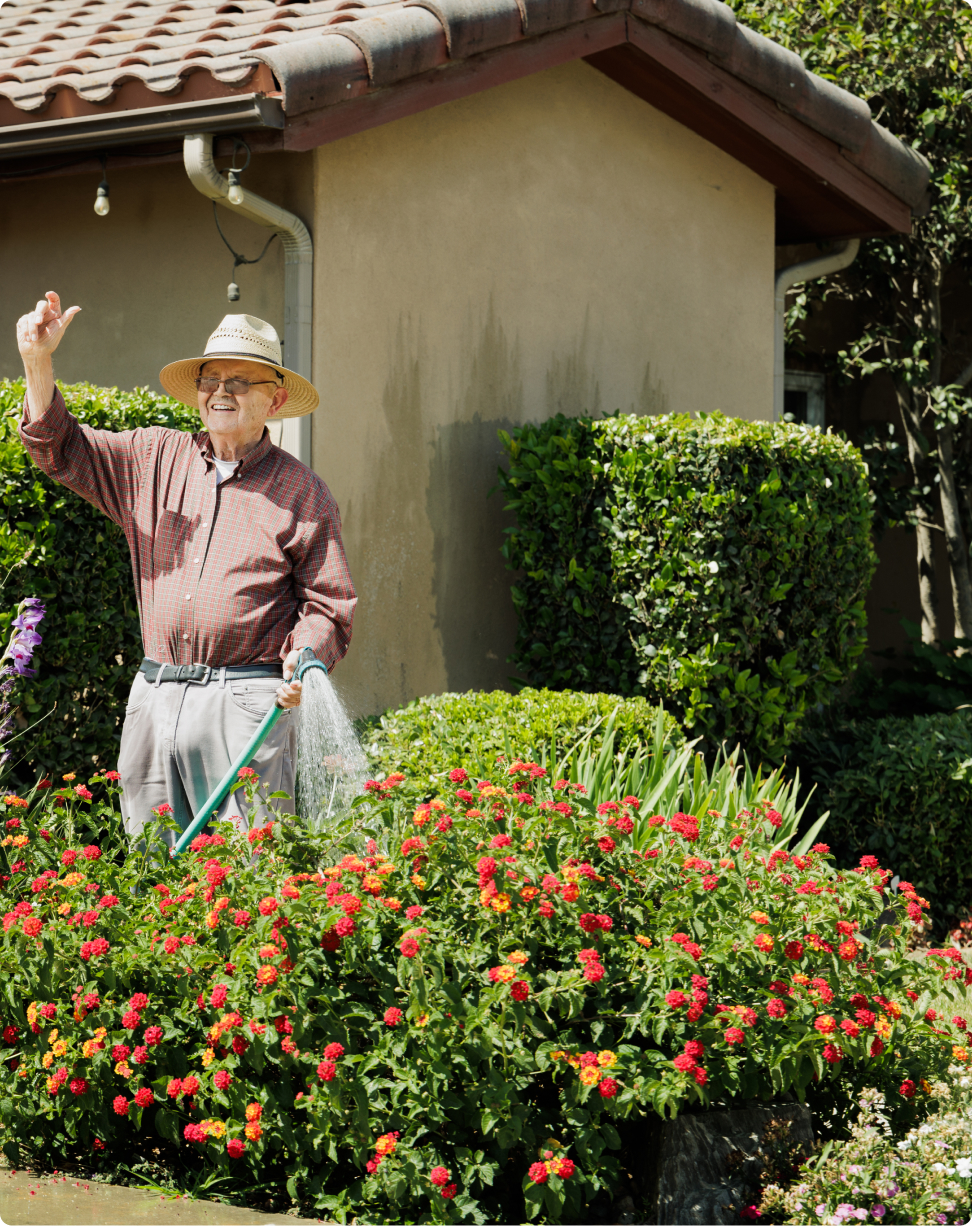 A person in a straw hat waters colorful garden flowers, standing by a house with a tiled roof, surrounded by greenery.