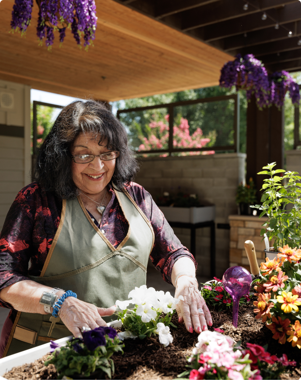 A person smiles while gardening, surrounded by colorful flowers and a wooden pergola. Bright sunlight illuminates the setting, creating a cheerful atmosphere.