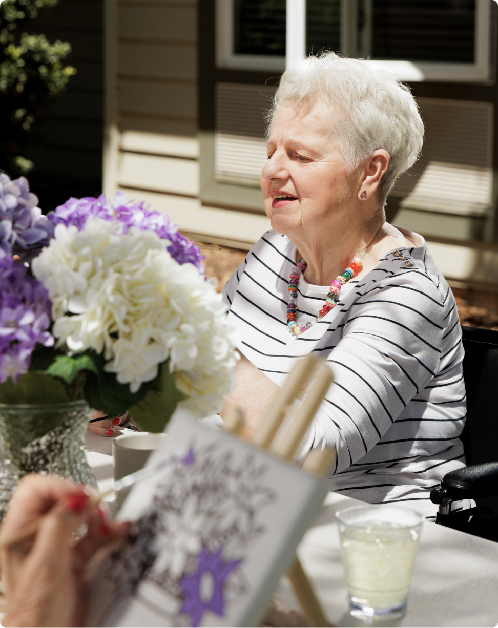 A person seated outdoors painting with flowers on the table, wearing a striped shirt and colorful necklace, enjoying a sunny day.