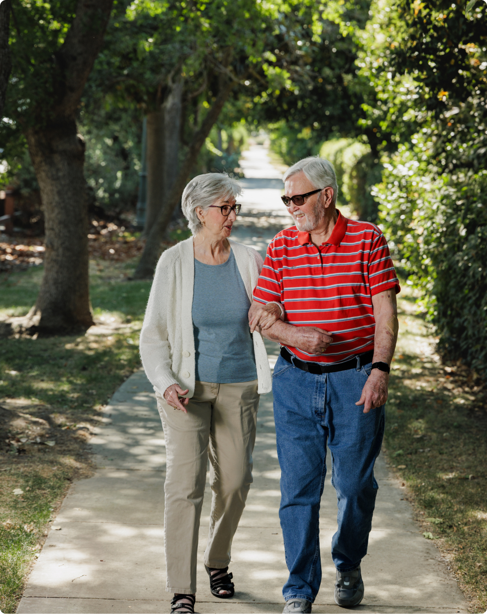 Two people walking arm-in-arm along a tree-lined path in a park, with sunlight filtering through the leaves, creating a peaceful atmosphere.