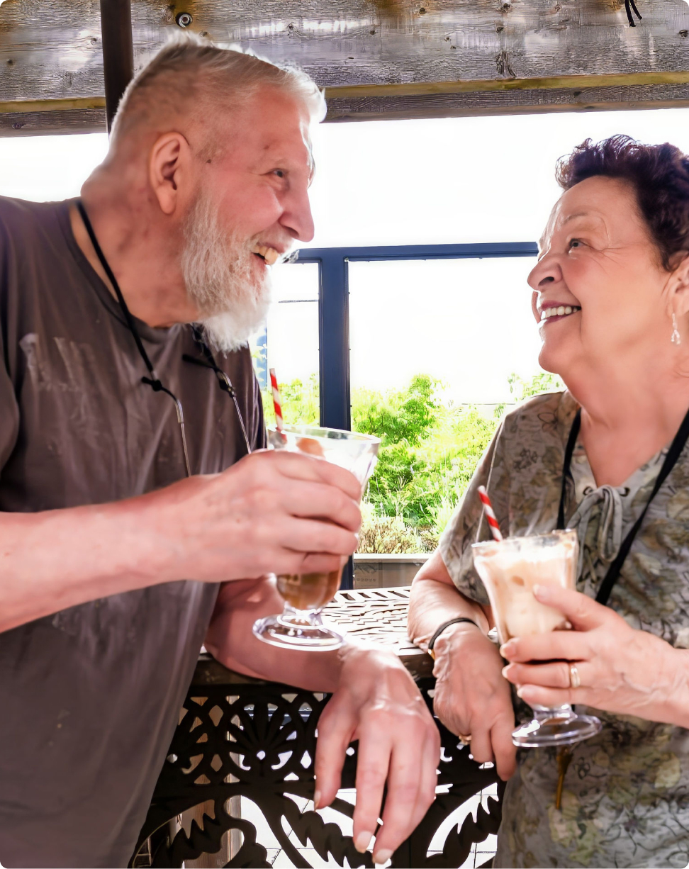 Two people smiling and enjoying drinks together on a balcony with a leafy green backdrop. Sunny weather and relaxed atmosphere.