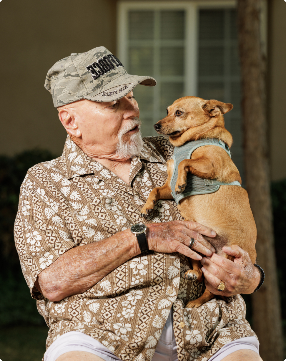 A person wearing a patterned shirt and cap holds a small dog, gazing affectionately, with a blurred building in the background.