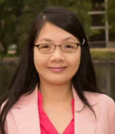 A person with glasses and long hair, wearing a pink jacket, stands outdoors with greenery in the background.