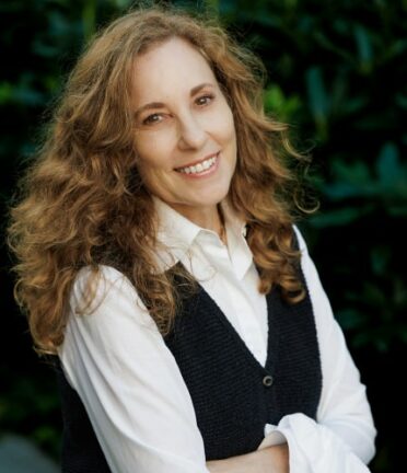 A person wearing a white long-sleeve shirt and vest smiling and standing against a leafy green background.