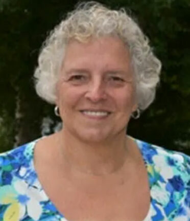 A person with short white hair, earrings and a blue floral shirt smiles directly at the camera.