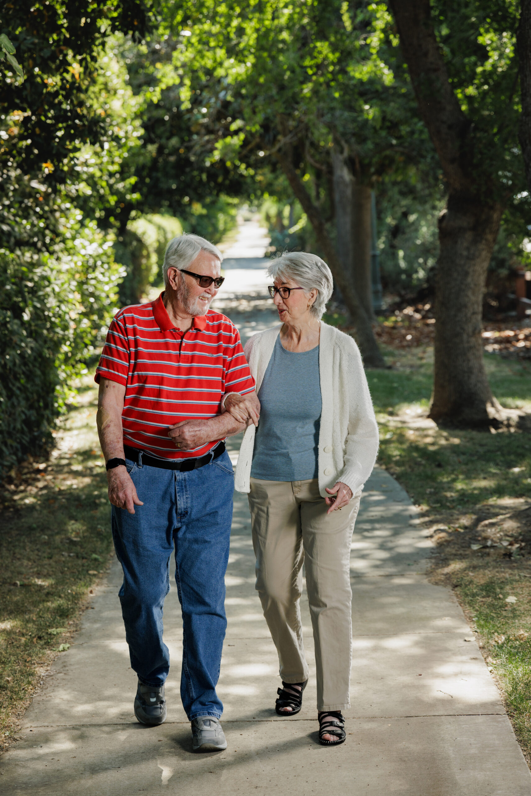 Two people walk arm in arm on a shaded path, surrounded by lush greenery and trees on a sunny day.