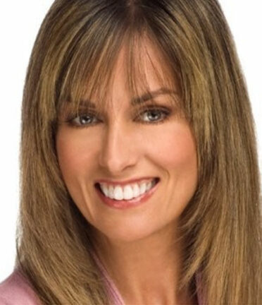 A person with brown hair and bangs smiles against a white background. The image is closely cropped, showing neck and head only.