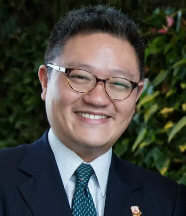 A person with short black hair, glasses and a dark suit and tie smiles at the camera.