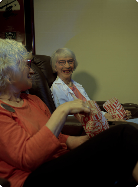 Two people sitting on a couch, smiling and eating popcorn. A "Miss Saigon" poster hangs on the wall in the background.