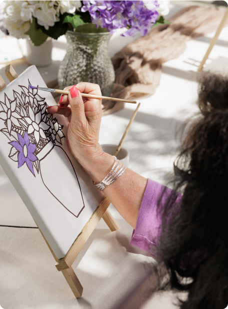 A person paints a flower vase on canvas, with purple flowers, in a bright setting. Decorative flowers are in the background.